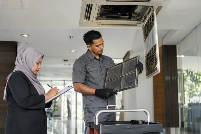 Juruteknik menyemak penapis penyaman udara sambil penyelia merekod servis.